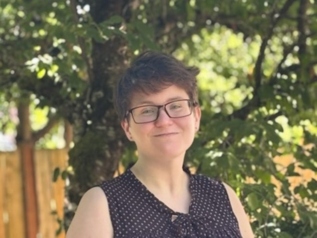 katie white headshot | Empowered Scholars Woman with short hair and glasses smiling outdoors in a sunny garden, wearing a black sleeveless blouse with white polka dots.
