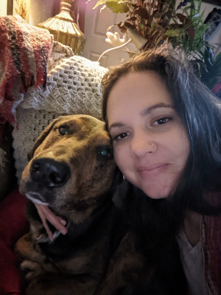 Sarah | Empowered Scholars Smiling woman taking a close-up selfie with a brown dog indoors, behind cozy decor and plants.